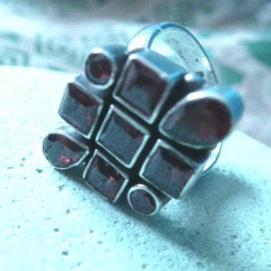 Puede incluir: Un anillo de plata con un diseño geométrico que presenta múltiples piedras preciosas de color rojo oscuro.
