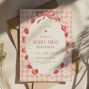 Puede incluir: Una invitación con estampado de cuadros rosas y tema de fresas. El texto dice "Emma's Berry First Birthday" con detalles del evento. El diseño incluye fresas, margaritas y un lazo rosa. La invitación está dentro de un sobre marrón.