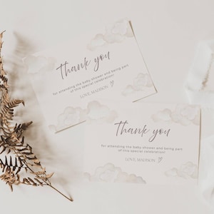 Może przedstawiać: Dwie karty z podziękowaniami na baby shower z ilustracjami chmur. Karty zawierają napis "Thank you" w ozdobnej czcionce, wraz z wiadomością i imieniem "LOVE, MADISON". Na zdjęciu znajduje się również suszona paproć i biała wstążka.