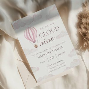 Peut inclure: Invitation de baby shower avec une illustration de montgolfière. L'invitation indique « We're on Cloud Nine » et comprend les détails de la baby shower de Madison Taylor. Le design présente une palette de couleurs douces avec des tons roses et beiges.