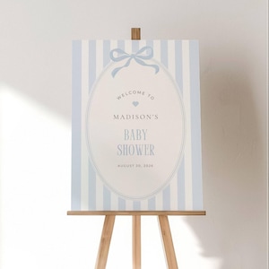 May include: A baby shower welcome sign on a wooden easel. The sign has a light blue and white striped background with an oval frame. The text reads "Welcome to Madison's Baby Shower August 20, 2026."