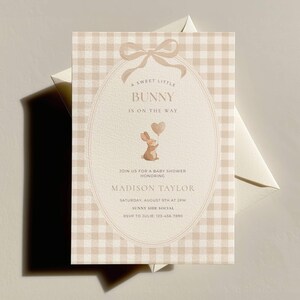 May include: Baby shower invitation with a beige gingham pattern background. The invitation features an oval design with a bow at the top and a watercolor illustration of a bunny holding a heart-shaped balloon. Text reads "A Sweet Little Bunny is on the Way."