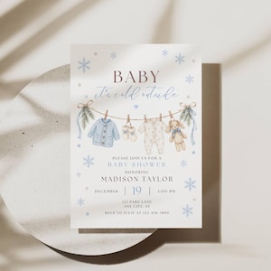 May include: A white baby shower invitation card featuring blue snowflakes and a clothesline with a blue cardigan, baby booties, a white onesie, and a teddy bear. The text reads Baby it's cold outside, Please join us for a baby shower honoring Madison Taylor, December 19, 1:00 PM, 123 Park Lane, Any City, ST, RSVP to Julie at 123-456-7890.