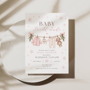 May include: A white baby shower invitation card featuring the text BABY it's cold outside, with a watercolor illustration of pink baby clothes and a teddy bear hanging on a clothesline, accented by pink snowflakes and event details.