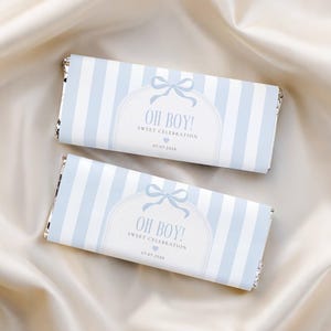 May include: Two chocolate bars wrapped in light blue and white striped paper. The wrappers feature the text "OH BOY! SWEET CELEBRATION" with a bow and the date 07.07.2028. The bars rest on a cream-colored satin surface.
