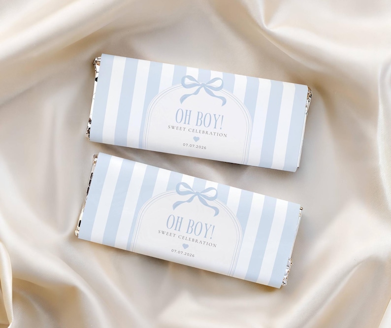 May include: Two chocolate bars wrapped in light blue and white striped paper. The wrappers feature the text "OH BOY! SWEET CELEBRATION" with a bow and the date 07.07.2028. The bars rest on a cream-colored satin surface.