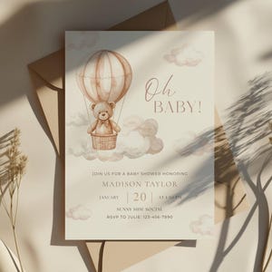 Può includere: Un invito per un baby shower con un design ad acquerello. L'invito presenta un orsacchiotto in mongolfiera, nuvole e il testo "Oh BABY!". L'invito è su uno sfondo color crema con una busta marrone.