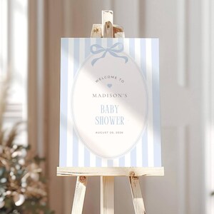 May include: A baby shower welcome sign on a wooden easel. The sign features a light blue and white striped background with an oval frame. The text reads "Welcome to Madison's Baby Shower August 20, 2026."