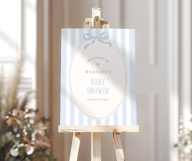May include: A baby shower welcome sign on a wooden easel. The sign features a light blue and white striped background with an oval frame. The text reads "Welcome to Madison's Baby Shower August 20, 2026."