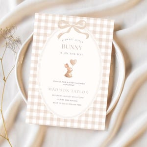 May include: A baby shower invitation with a beige gingham pattern background. The invitation features a bunny holding a heart-shaped balloon and the text "A Sweet Little Bunny Is On The Way". Details include the honoree's name, date, time, and RSVP information.