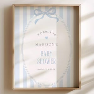 May include: A framed baby shower sign with a light blue and white striped background. The sign features an oval design with the words "Welcome to Madison's Baby Shower" and the date August 20, 2026, in light blue lettering.