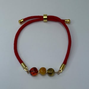 May include: Red adjustable bracelet with three amber beads in shades of orange, yellow, and green. The bracelet features gold-colored metal accents and a sliding knot closure for a custom fit. A stylish accessory.