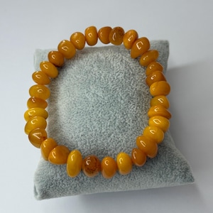 May include: A bracelet made of polished amber beads, each with a unique shape and a warm, golden hue. The beads are strung together to form a circular bracelet. The bracelet is displayed on a light grey cushion.