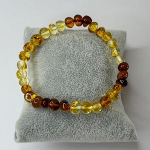 May include: A bracelet made of amber beads, displaying a range of yellow and brown hues. The beads are round and polished, strung together to form the bracelet. The piece is presented on a grey surface, showcasing its natural tones.