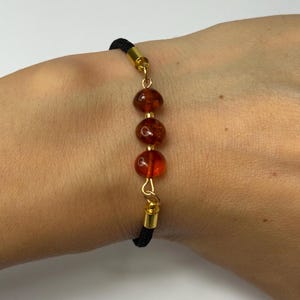 May include: A bracelet with three amber beads on a black cord. The beads are linked with gold-coloured metal. The bracelet is designed to be worn on the wrist.