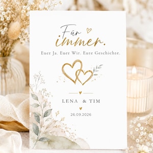 Könnte beinhalten: Eine rechteckige weiße Hochzeitseinladungskarte mit goldener Schrift "Für immer. Euer Ja. Euer Wir. Eure Geschichte.", zwei ineinander verschlungenen goldenen Herzen, grünen Aquarellblättern sowie den Namen "LENA & TIM" und dem Datum "26.09.2026".
