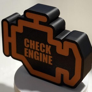May include: A black and orange 3D-printed object in the shape of a car engine warning light. The text "CHECK ENGINE" is printed in white within the orange outline. The object is sitting on a white surface.