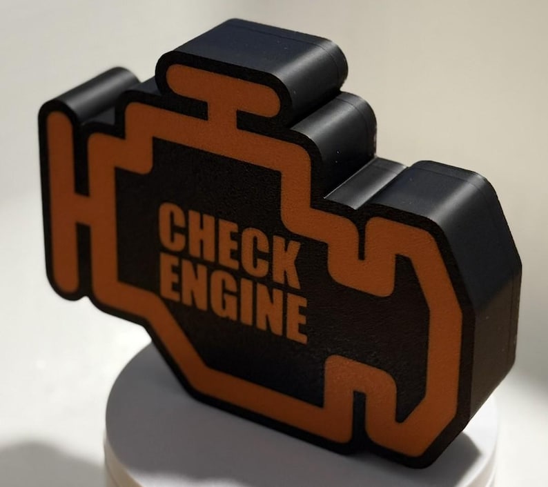 May include: A black and orange 3D-printed object in the shape of a car engine warning light. The text "CHECK ENGINE" is printed in white within the orange outline. The object is sitting on a white surface.
