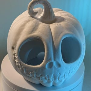 May include: A white, unpainted pumpkin-shaped decorative item with large, oval eye holes and a stitched-mouth design. The pumpkin has a curly stem and is likely made of ceramic or a similar material. The design is reminiscent of a jack-o'-lantern.
