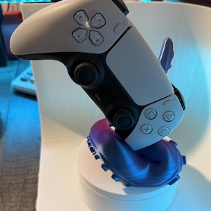 May include: A white and black video game controller resting on a purple and blue stand. The stand has a unique, curved design. The controller features multiple buttons and directional controls. The stand is on a white, circular base.
