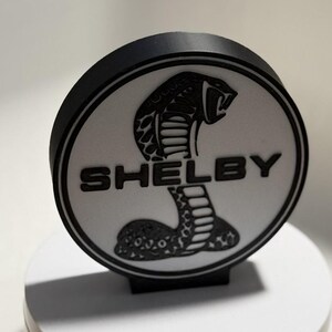 Op de afbeelding: Een zwart-wit Shelby-embleem met een cobra-motief. Het ronde embleem heeft het woord "SHELBY" in vette letters. De cobra staat centraal, met zijn kop omhoog en bek open.