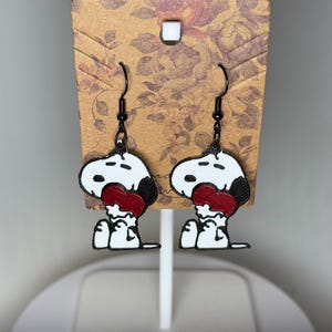 May include: A pair of Snoopy earrings. Each earring features a white Snoopy head with black accents and a red heart-shaped tongue. The earrings hang from black fish hook ear wires and are displayed on a floral patterned card.