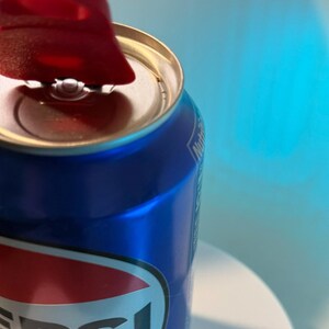 May include: Close-up of a blue Pepsi can with a red and white logo. The can's pull tab is open, revealing the silver interior. The background is a gradient of blue and white.