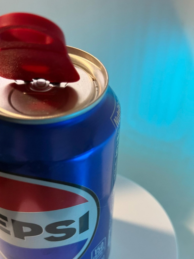 May include: Close-up of a blue Pepsi can with a red and white logo. The can's pull tab is open, revealing the silver interior. The background is a gradient of blue and white.