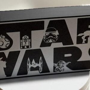 Könnte beinhalten: Schwarzes und silbernes Star Wars Logo mit Silhouetten von Charakteren. Das Logo zeigt den Text "STAR WARS" und Bilder von BB-8, R2-D2, einem Sturmtruppler, Yoda und anderen ikonischen Charakteren.