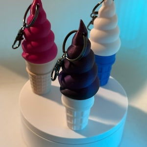 Puede incluir: Tres llaveros de cono de helado en rosa, morado y blanco. Los conos de helado tienen un diseño en espiral y se asientan sobre bases de cono de gofre. Cada llavero tiene un anillo y un cierre plateados. Los conos se exhiben en una plataforma redonda blanca.