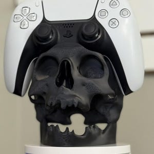 Puede incluir: Una escultura de calavera negra con un mando de PlayStation 5 blanco y negro encima. El mando presenta la disposición estándar de los botones y un logotipo de PlayStation. La calavera es detallada y se asienta sobre una base blanca.