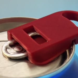 May include: A close-up of a red can opener attached to a silver soda can. The can opener has a rectangular design with a loop for easy handling. The can is blue and silver.