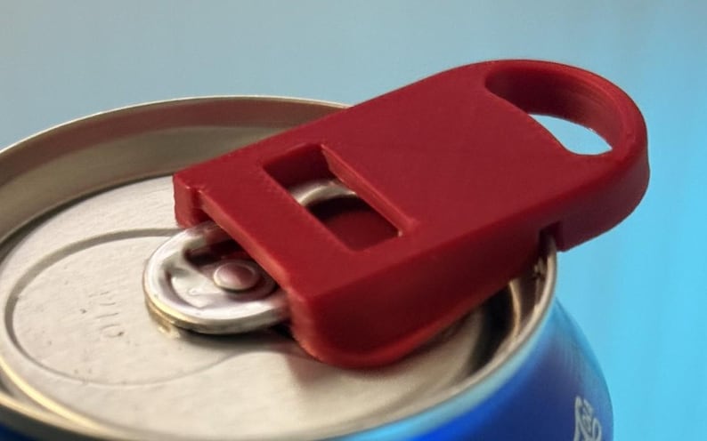 May include: A close-up of a red can opener attached to a silver soda can. The can opener has a rectangular design with a loop for easy handling. The can is blue and silver.