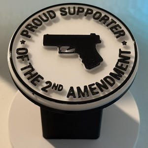 Puede incluir: Un objeto redondo blanco y negro con el texto "PROUD SUPPORTER OF THE 2ND AMENDMENT" que rodea una silueta de pistola negra. El objeto tiene una base negra.