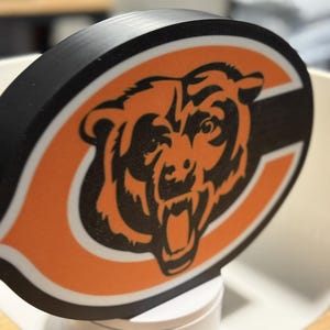 May include: A black and orange Chicago Bears logo, featuring a bear's head within a white and black 'C' shape. The logo is set against a white background. The bear is orange with black details.
