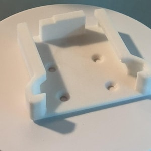 May include: A white 3D-printed plastic bracket with a rectangular base and two vertical supports. The bracket has several holes and is designed to hold an object. The supports have a curved shape.