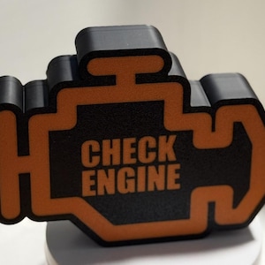 May include: A black and orange 3D-printed check engine light. The words "CHECK ENGINE" are printed in orange on the black surface. The object is on a white surface. The design is a common automotive symbol.