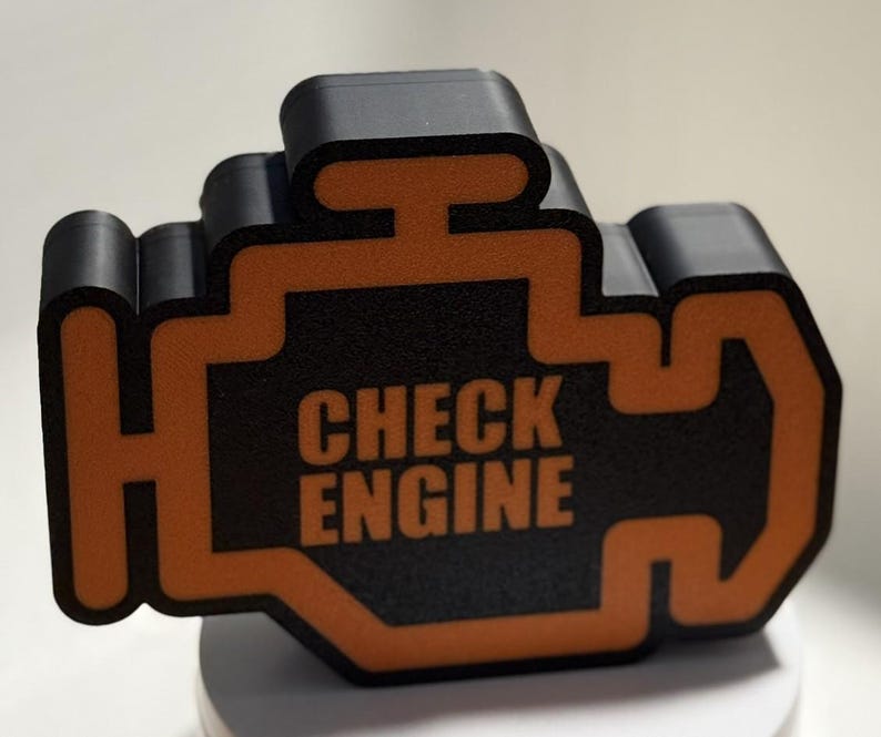 May include: A black and orange 3D-printed object in the shape of a car's check engine light. The words "CHECK ENGINE" are printed in orange on the black surface. The object is sitting on a white surface.