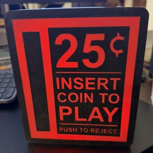 Può includere: Un cartello rettangolare nero e rosso con il testo 25 cents, INSERT COIN TO PLAY e PUSH TO REJECT. Il design utilizza lettere rosse in grassetto su sfondo nero, con una barra verticale rossa sul lato sinistro del blocco di testo.