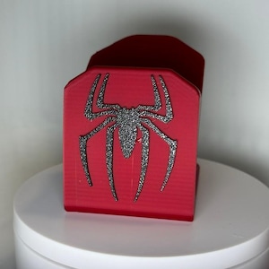 Puede incluir: Un tarjetero rojo rectangular con un diseño de araña negro brillante. El tarjetero está hecho de un material sólido y se muestra sobre una superficie blanca. El diseño de la araña está centrado en la parte delantera.