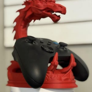 May include: A red dragon-shaped game controller stand holding a black controller. The dragon has a detailed, textured design and is made of a solid material. The controller is resting in the dragon's claws.