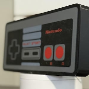 May include: A black and grey Nintendo controller with red buttons and the brand name in red. The controller features a directional pad, start and select buttons, and A and B buttons. This is a retro gaming item.
