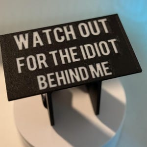 May include: A black sign with white text that reads "WATCH OUT FOR THE IDIOT BEHIND ME." The sign is mounted on a small stand. The background is a gradient of white and blue.