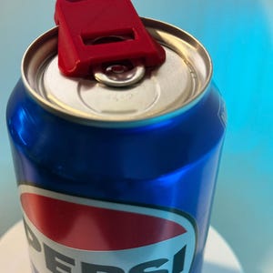 May include: A blue Pepsi can with a red and white logo, topped with a red plastic can opener. The can opener is designed to lift the tab for easy opening. The background is a gradient of blue.