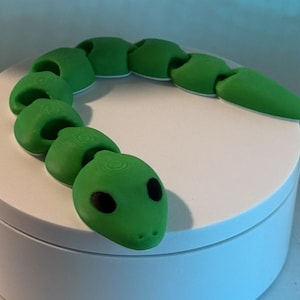 Peut inclure: Un jouet serpent articulé vert avec une tête arrondie et un corps segmenté. Le serpent a des yeux noirs et une base blanche. Le jouet est en plastique et est conçu pour être flexible et mobile.