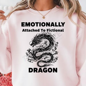 Puede incluir: Una sudadera rosa claro con el texto "Emotionally Attached To Fictional Dragon" en negro. Un detallado gráfico de dragón en blanco y negro está centrado debajo del texto. El dragón está rodeado de líneas y formas arremolinadas.