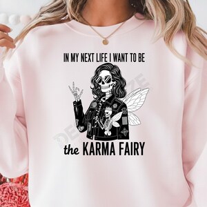 Puede incluir: Sudadera rosa claro con el texto "IN MY NEXT LIFE I WANT TO BE the KARMA FAIRY". El gráfico es una ilustración en blanco y negro de un hada esqueleto con gafas de sol y una chaqueta negra.