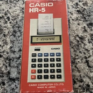 May include: A vintage Casio HR-5 printing calculator in its original box. The red box features the Casio logo and model number. The calculator has a white body with black keys and a digital display showing the number 1234567890. Made in Japan.