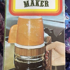 May include: Vintage "Auto Coffee Maker" box featuring an orange coffee cup and a glass carafe. The box has text that reads "Auto Coffee Maker" and "Just plug into 12-volt cigarette lighter socket."
