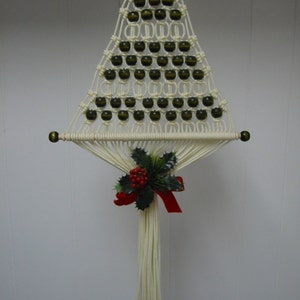 May include: A macrame Christmas tree wall hanging. The off-white tree is adorned with dark green beads and a holly and berry accent with a red ribbon. The tree is suspended from a gold ring and has a fringe at the bottom.